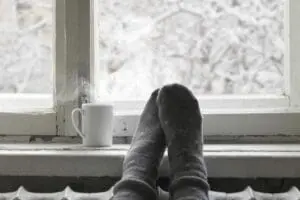 How Do I Keep My Heat During Extreme Cold Weather? Image shows socked feet resting on windowsill next to hot drink.