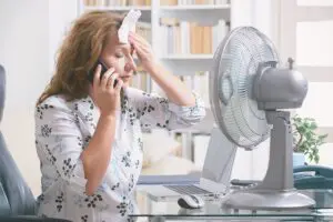 When Do I Replace My Air Conditioner? Woman on the phone attempting to cool herself with a fan.
