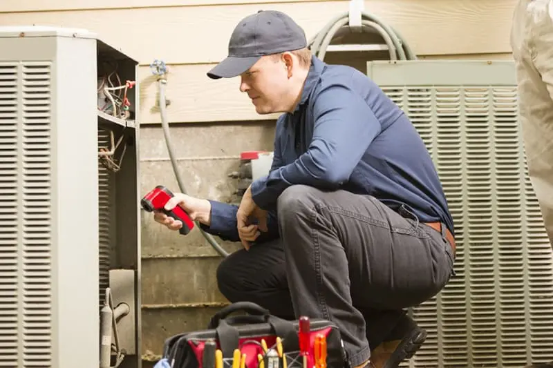 Blogs - Just Right Learn About Our No-Breakdown Guarantee - Mid-adult repairman works on a home's air conditioner unit outdoors. Man center is working on the unit using tools in his toolbox. Other man to right. They both wear uniforms.