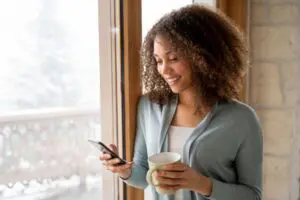 lady looking at her phone, holding a coffee cup and standing beside a window