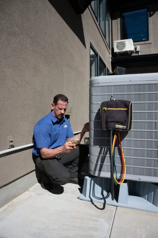 HVAC technician performing AC maintenance in Layton UT beside an outdoor residential air conditioning condenser unit during a cooling system inspection. The image shows the technician using HVAC diagnostic tools and refrigerant hoses connected to the condenser while completing seasonal maintenance service. Just Right Plumbing, Heating & Cooling provides AC maintenance, cooling system inspections, and residential HVAC tune-ups for homeowners throughout Layton, Utah. Customers needing AC maintenance in Layton UT can contact Just Right Plumbing, Heating & Cooling at (801) 771-1510 for cooling system service, condenser inspections, and preventative HVAC maintenance.