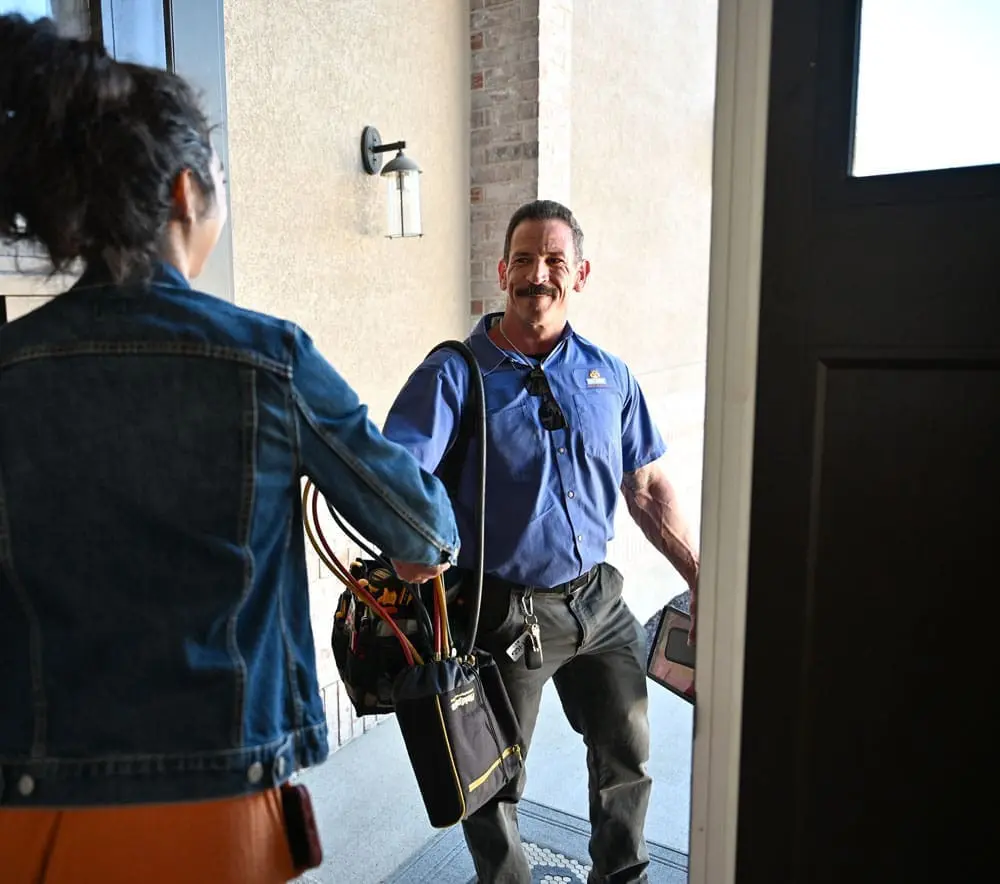 HVAC technician arriving for AC services in Bluffdale UT while greeting a homeowner at the front entrance of a residential home. The image shows a Just Right Plumbing, Heating & Cooling technician carrying HVAC service tools and refrigerant hoses during a residential cooling system appointment. The visible service equipment, technician uniform, and homeowner interaction reflect real residential AC service visits focused on professional customer care and indoor comfort support throughout Bluffdale, Utah.