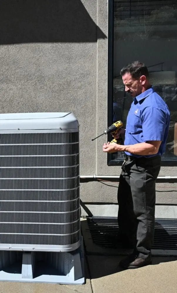 HVAC technician performing AC services in Layton UT beside an outdoor air conditioning condenser unit during a residential cooling system maintenance visit. The image shows a service technician holding repair tools while inspecting an exterior AC system near a home. Just Right Plumbing, Heating & Cooling provides air conditioning repair, AC maintenance, and cooling system inspections for homeowners throughout Layton, Utah. Customers needing AC services in Layton UT can call Just Right Plumbing, Heating & Cooling at (801) 771-1510 for same-day cooling repairs, seasonal tune-ups, and HVAC diagnostics.