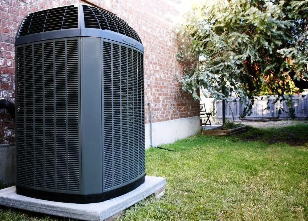 A high-efficiency outdoor air conditioning unit is installed beside a brick home in Bluffdale, UT. Just Right Plumbing, Heating & Cooling provides AC installation and replacement services for homeowners throughout Bluffdale and nearby Utah communities. The image shows a modern residential condenser system mounted on a concrete pad with a maintained lawn and shaded backyard environment.