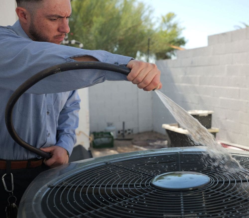 AC Maintenance Alpine UT service from Just Right Plumbing, Heating & Cooling shows a technician cleaning an outdoor air conditioning condenser with a water hose at a residential property. The image includes a close-up view of the AC fan grille, water spray, and outdoor cooling equipment during seasonal maintenance service. Just Right Plumbing, Heating & Cooling provides AC maintenance, cooling inspections, and preventive HVAC service for homeowners in Alpine, UT. Call (801) 302-1154 to schedule AC Maintenance Alpine UT service with a local HVAC technician.
