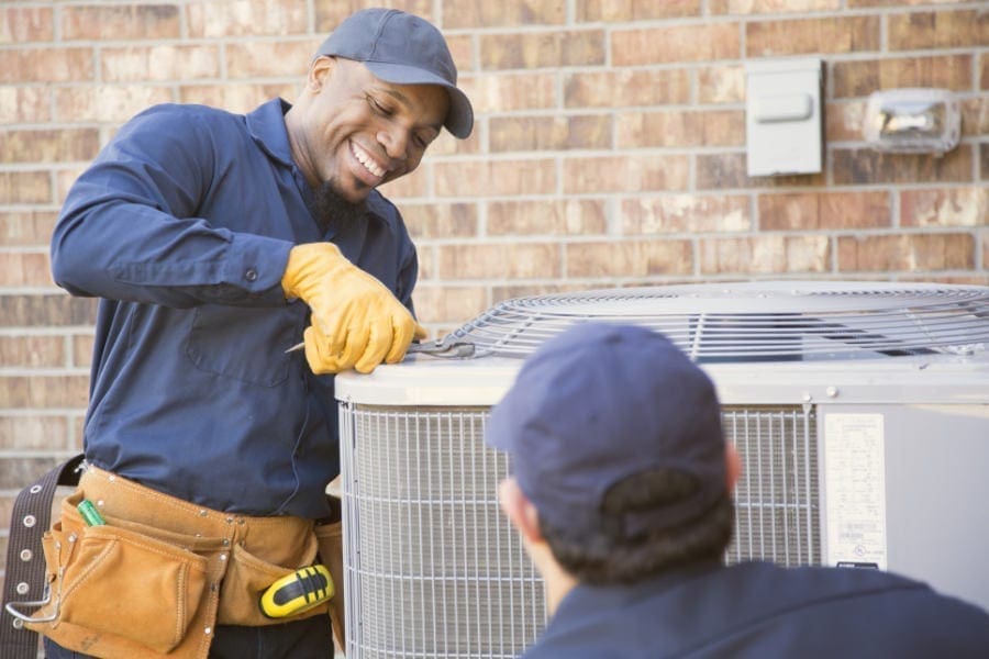 AC Maintenance American Fork UT service from Just Right Plumbing, Heating & Cooling shows two HVAC technicians working on an outdoor air conditioning condenser unit beside a residential brick wall. The image includes technicians using hand tools, work gloves, and a tool belt while performing cooling system maintenance on a home AC unit. Just Right Plumbing, Heating & Cooling provides AC maintenance, cooling inspections, and seasonal HVAC service for homeowners in American Fork, UT. Call (801) 302-1154 for AC Maintenance American Fork UT service from a local HVAC team.