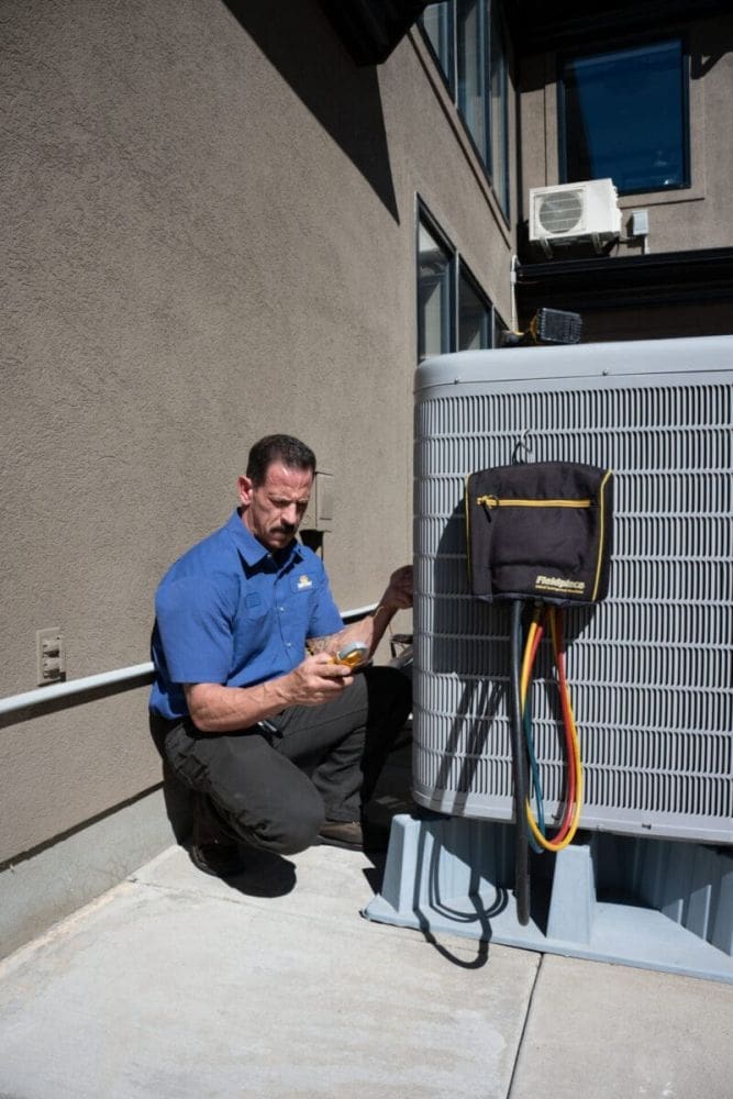 HVAC technician performing AC maintenance in Layton UT beside an outdoor residential air conditioning condenser unit during a cooling system inspection. The image shows the technician using HVAC diagnostic tools and refrigerant hoses connected to the condenser while completing seasonal maintenance service. Just Right Plumbing, Heating & Cooling provides AC maintenance, cooling system inspections, and residential HVAC tune-ups for homeowners throughout Layton, Utah. Customers needing AC maintenance in Layton UT can contact Just Right Plumbing, Heating & Cooling at (801) 771-1510 for cooling system service, condenser inspections, and preventative HVAC maintenance.