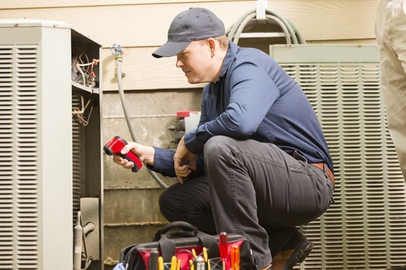 HVAC technician performing AC maintenance in South Salt Lake UT while inspecting an outdoor residential air conditioning condenser unit with a handheld diagnostic device. The image shows cooling system maintenance work beside an open condenser panel during a residential HVAC service appointment. Just Right Plumbing, Heating & Cooling provides AC maintenance, cooling system inspections, and residential HVAC tune-ups for homeowners in South Salt Lake, Utah.