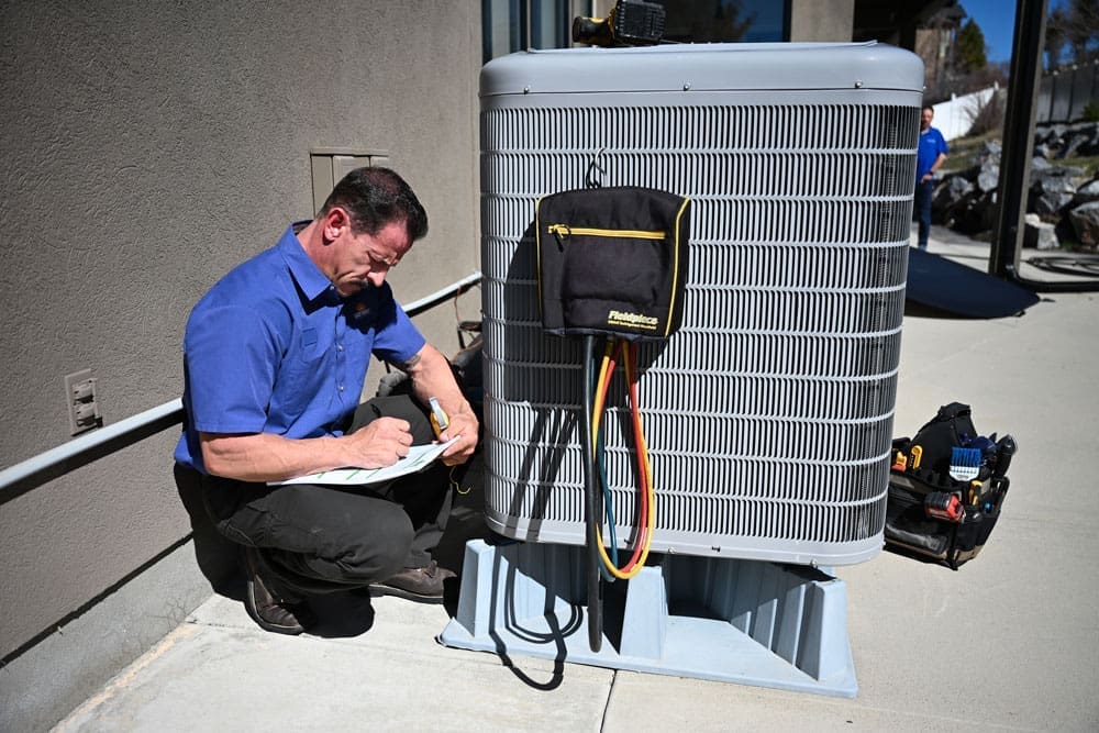 HVAC technician performing AC services in Bountiful UT while completing cooling system inspection paperwork beside an outdoor air conditioning condenser unit. The image shows a residential HVAC service appointment with refrigerant hoses, diagnostic equipment, and service tools connected to the condenser during maintenance work. Just Right Plumbing, Heating & Cooling provides AC services, cooling system inspections, and residential HVAC repairs for homeowners in Bountiful, Utah.