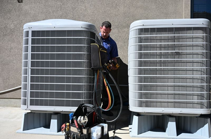 HVAC technician performing AC services in Layton UT beside two outdoor air conditioning condenser units during a cooling system inspection. Just Right Plumbing, Heating & Cooling provides air conditioning repair, AC maintenance, and cooling system service for homeowners in Layton, Utah. The image shows an HVAC worker using gauges and service tools while checking residential cooling equipment near a building exterior. Customers in Layton can call Just Right Plumbing, Heating & Cooling at (801) 771-1510 for same-day AC repair, air conditioning tune-ups, and cooling system diagnostics.