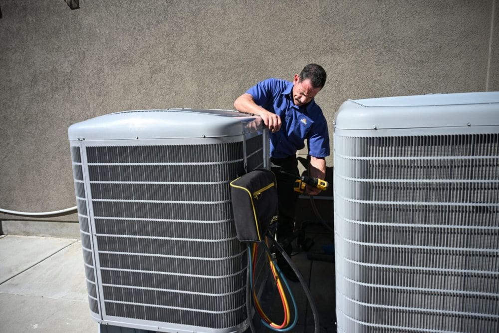 HVAC technician performing AC Installation and Replacement Alpine UT service on outdoor air conditioning condenser units at a residential property. The technician is inspecting and connecting cooling system equipment while using professional HVAC tools during an air conditioner installation project. This image represents AC replacement, AC installation, and cooling system setup services provided by Just Right Plumbing, Heating & Cooling in Alpine, Utah.