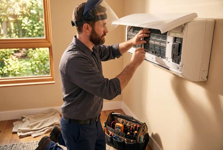 Bearded HVAC technician wearing a face shield working on a wall-mounted mini-split AC unit with a tool bag nearby in Layton UT