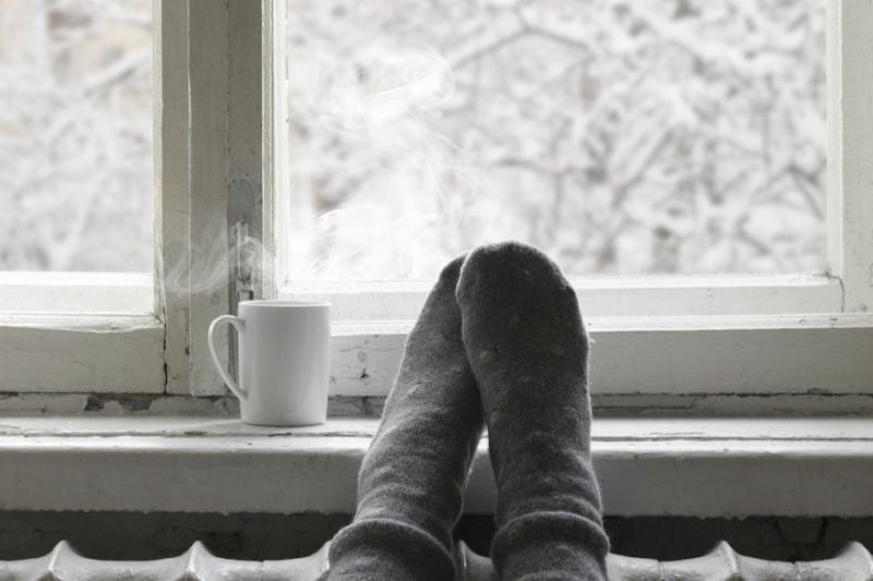 How Do I Keep My Heat During Extreme Cold Weather? Image shows socked feet resting on windowsill next to hot drink.