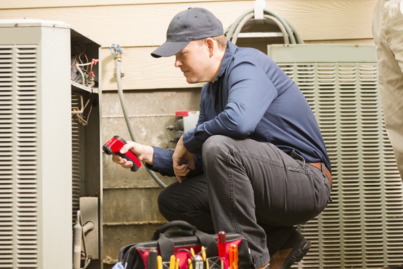 Learn About Our No-Breakdown Guarantee - Mid-adult repairman works on a home's air conditioner unit outdoors. Man center is working on the unit using tools in his toolbox. Other man to right. They both wear uniforms.