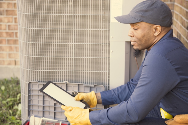 African descent, blue collar air conditioner repairman at work. He refers to digital tablet for next steps.